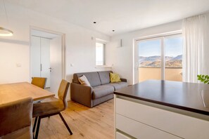A modern living room with a wooden dining table, a grey sofa, and a kitchen area with a large window offering a view of the mountains.