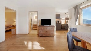 Una sala moderna con piso de madera, un televisor sobre un aparador de madera, una mesa de comedor con sillas y una zona de cocina al fondo.