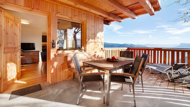 Una terraza de madera con una mesa y sillas de comedor, un televisor al fondo y vistas a las montañas.