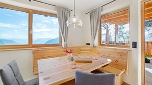Zona de estar con bancos de madera, una mesa, una lámpara colgante y ventanas grandes con cortinas.