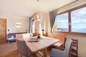 A dining area with a wooden table, chairs, a hanging light fixture, and a view through large windows.