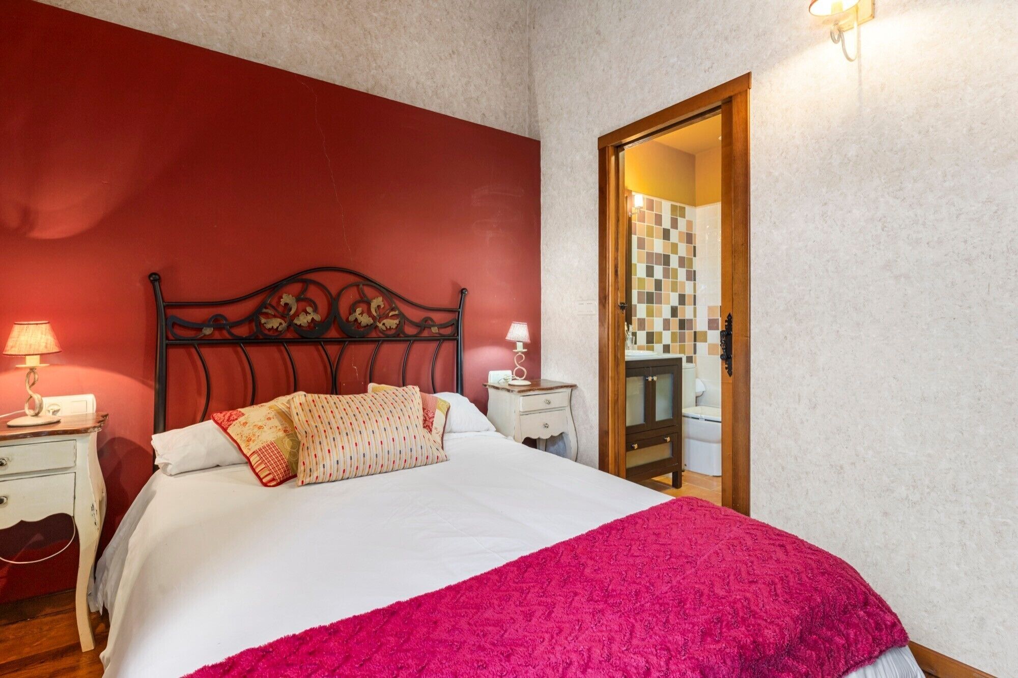 Un dormitorio con una cama, dos mesitas de noche, una mesita auxiliar, una puerta de madera y un baño visible a través de una puerta de cristal.