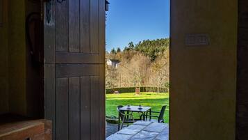 An open wooden door leading to a patio with a table and chairs, a small shed, and a grassy area with trees.