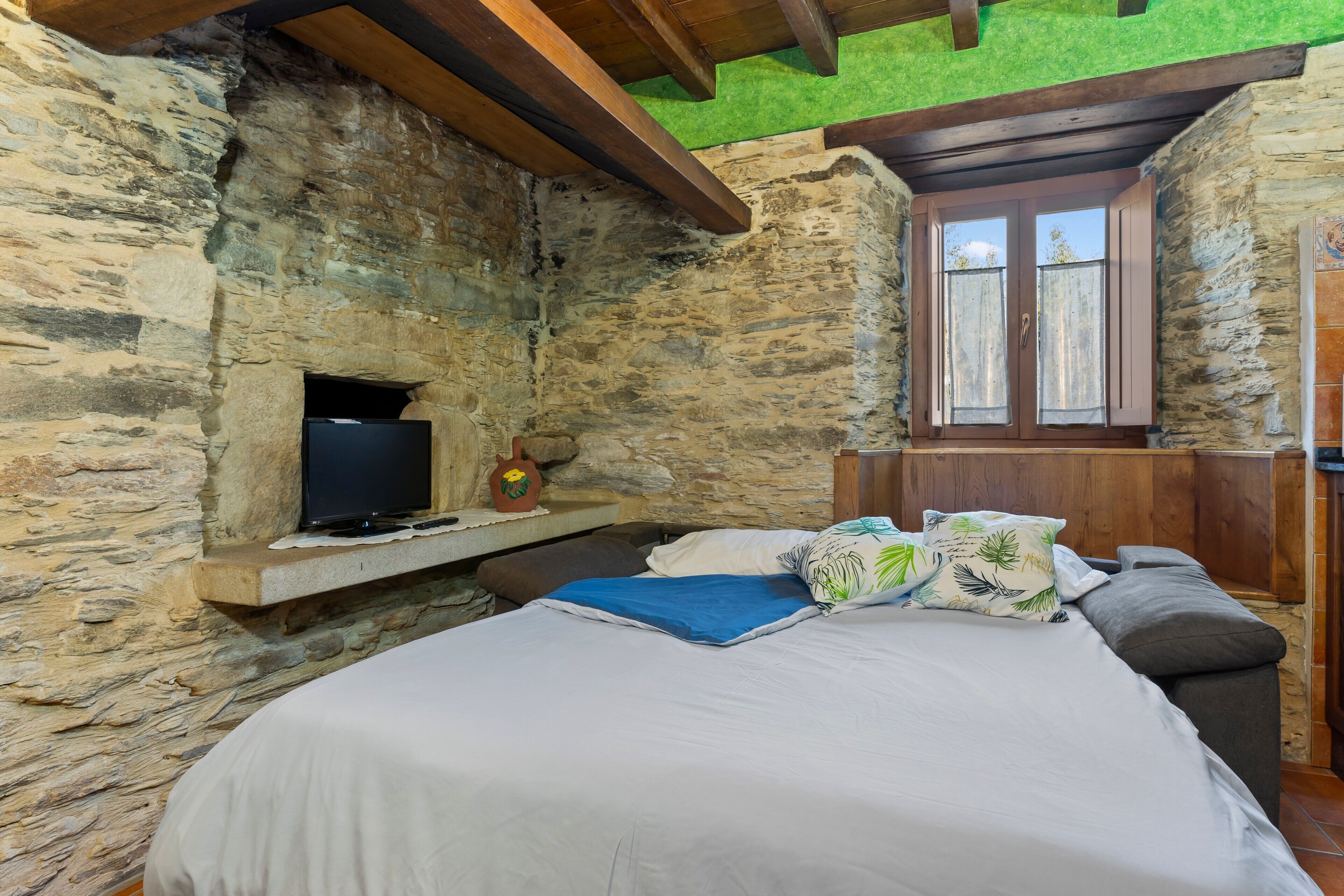 Un dormitorio con paredes de piedra, una cama, almohadas, un televisor y una ventana.