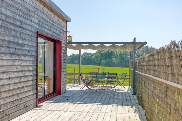 Une terrasse en bois avec une table et des chaises, une porte à cadre rouge et une vue sur un champ verdoyant.