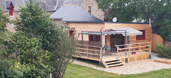 A wooden cabin with a deck, a small outdoor seating area, and a stone building in the background.