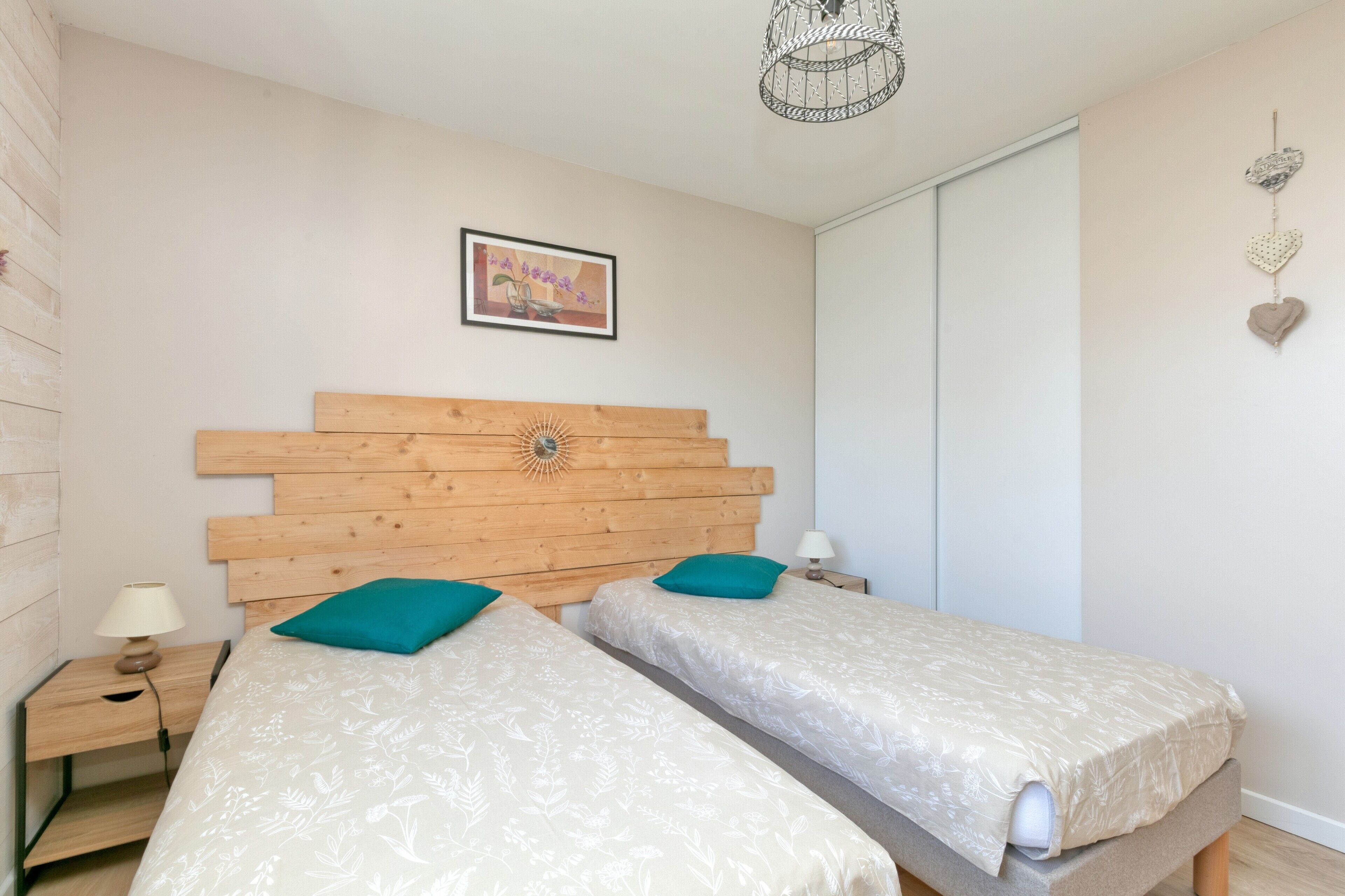 A bedroom with two single beds, a wooden headboard, a bedside table with a lamp, a framed picture on the wall, and a hanging heart decoration.