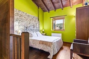 A bedroom with a stone wall, a bed with a patterned bedspread, a wooden chair, and a window with a view of trees.