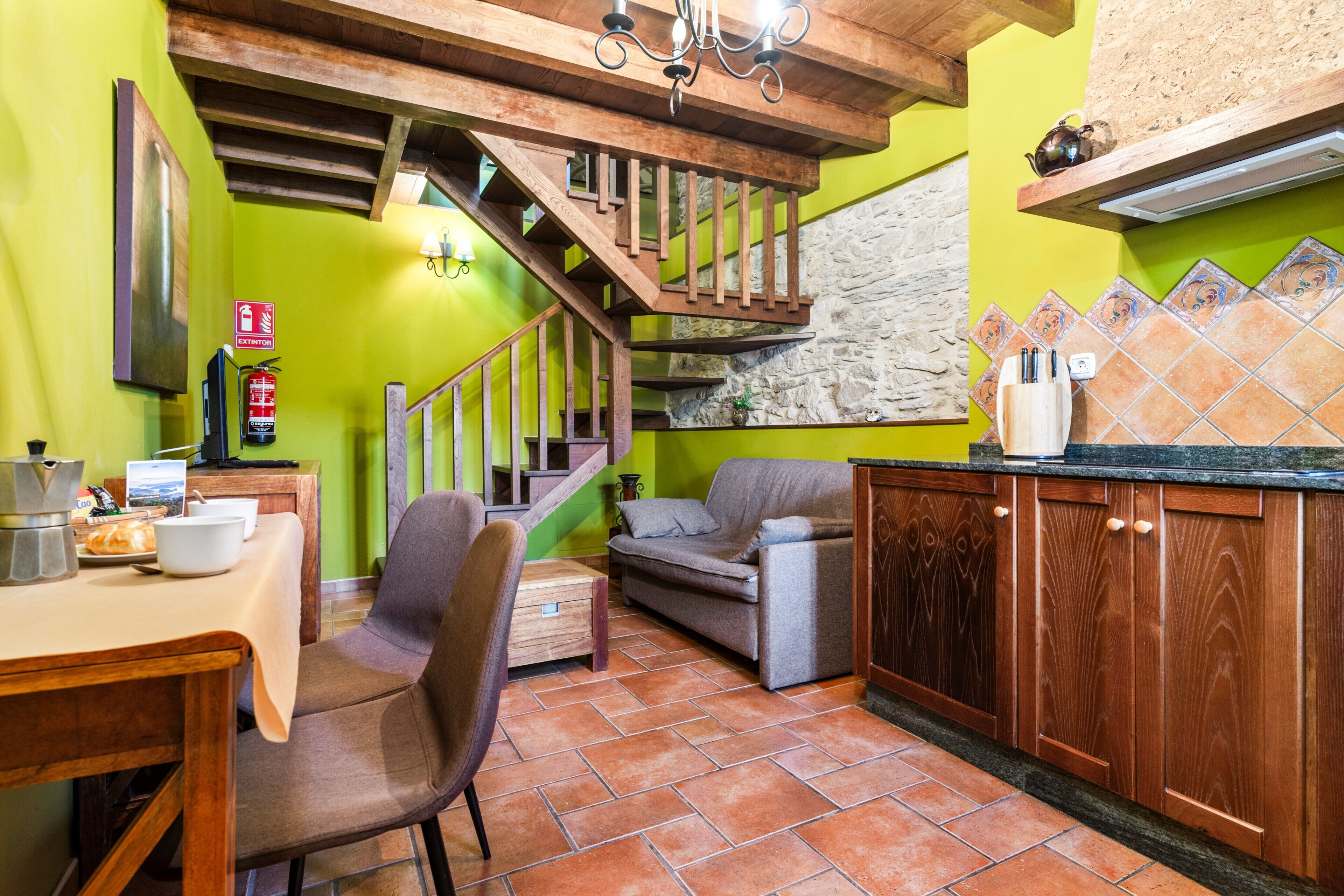 Une cuisine avec un escalier en bois, une table à manger avec des chaises, un canapé et un mur recouvert de carreaux.