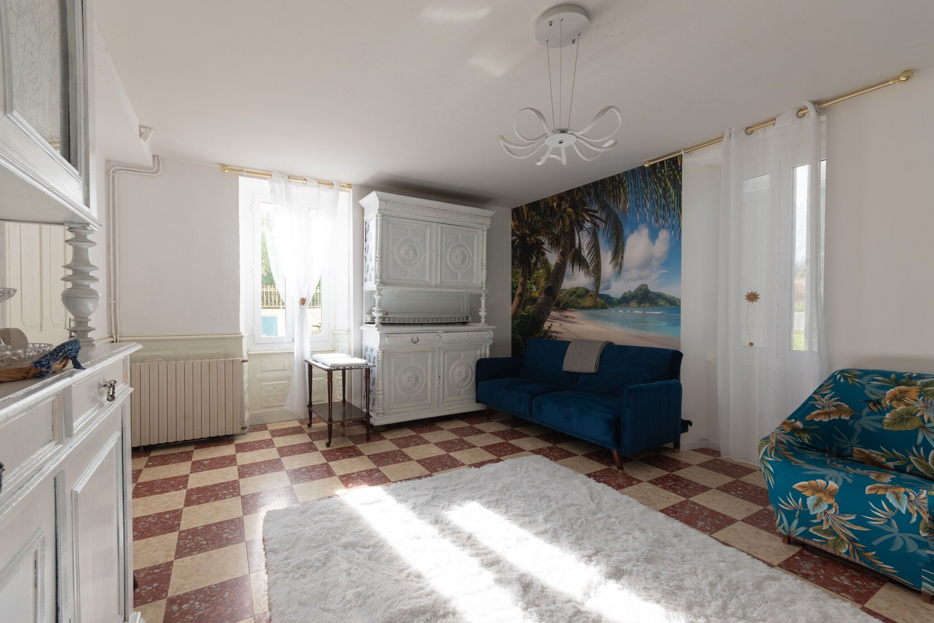 Une pièce avec un sol à carreaux, un radiateur blanc, un canapé bleu, une armoire blanche et un tableau représentant une scène de plage.