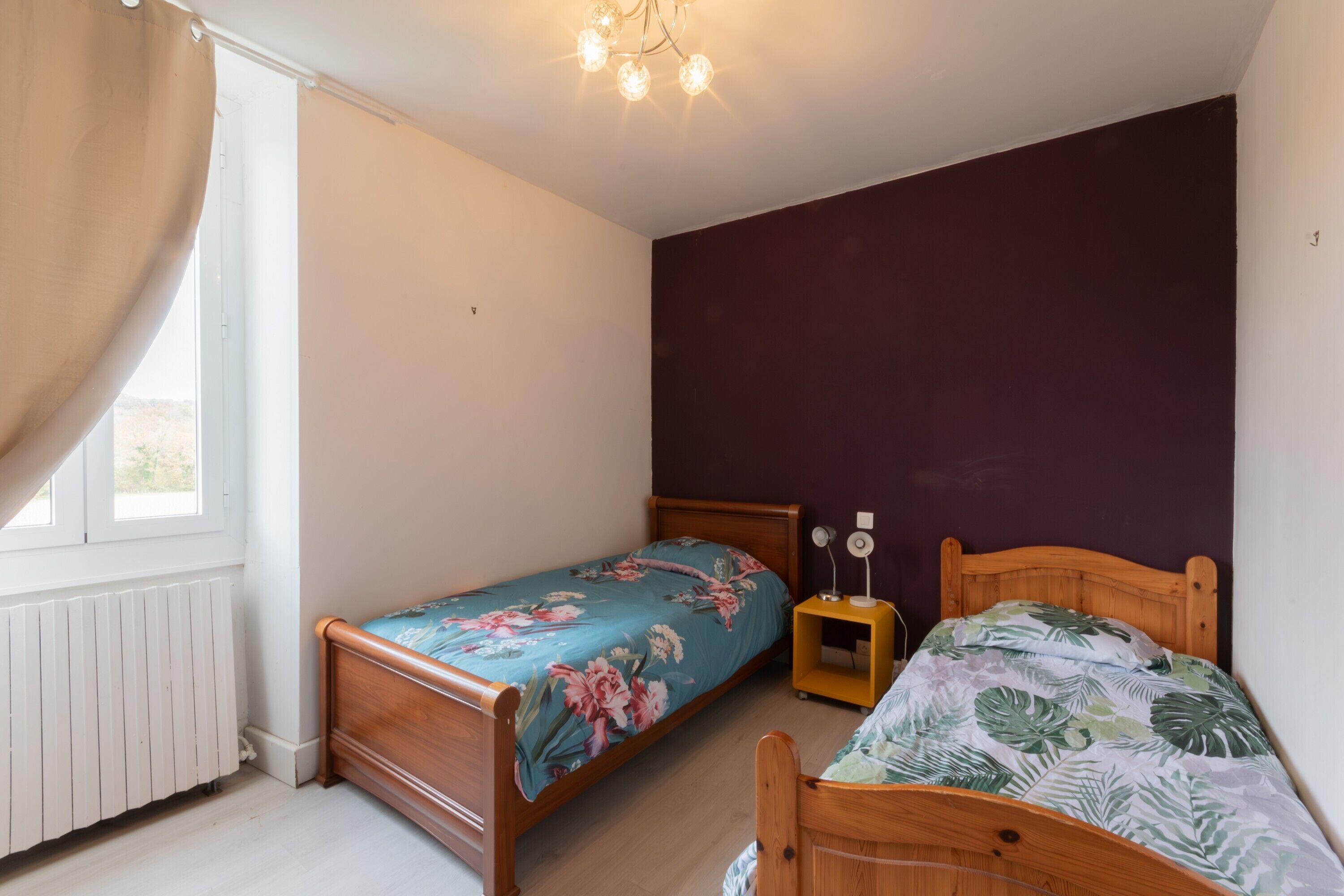 Une chambre avec deux lits en bois, une fenêtre avec des rideaux, un radiateur et un luminaire fixé au plafond.