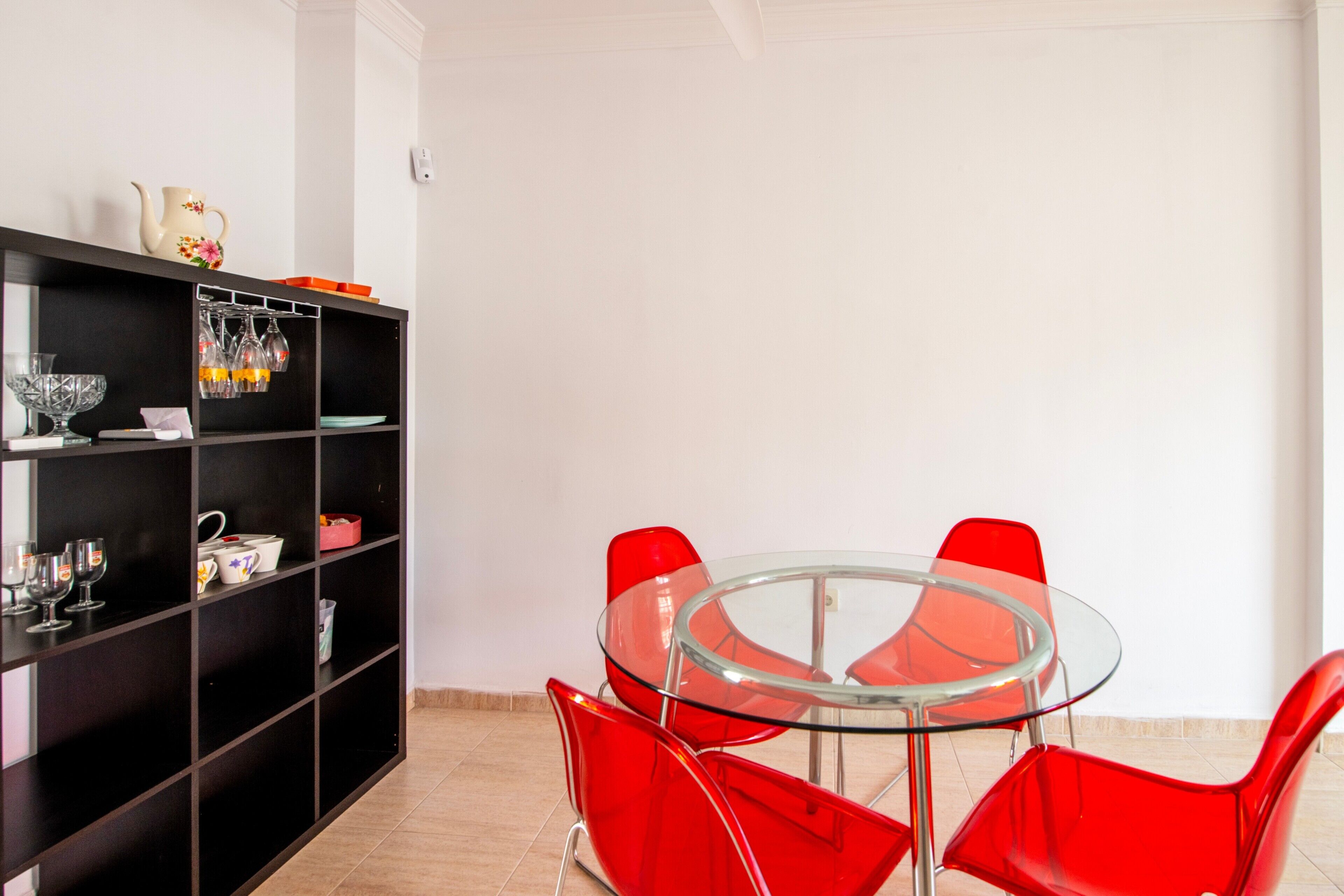 Een moderne eetruimte met een glazen tafel en rode stoelen, een zwart wandrek met keukengerei, en een witte muur.