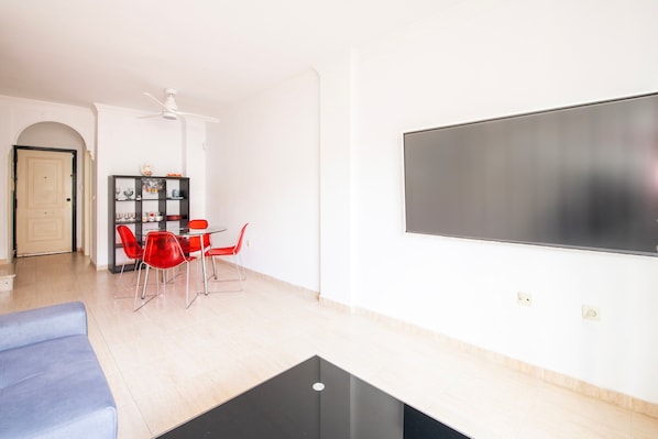 A living room with a black coffee table, a blue sofa, a dining area with red chairs, a white wall-mounted TV, and a ceiling fan.