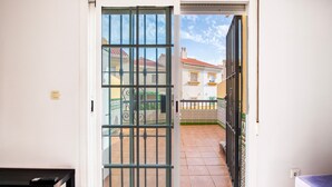Una puerta corrediza de vidrio da a un balcón con piso de baldosas, baranda de metal y vista de edificios vecinos.