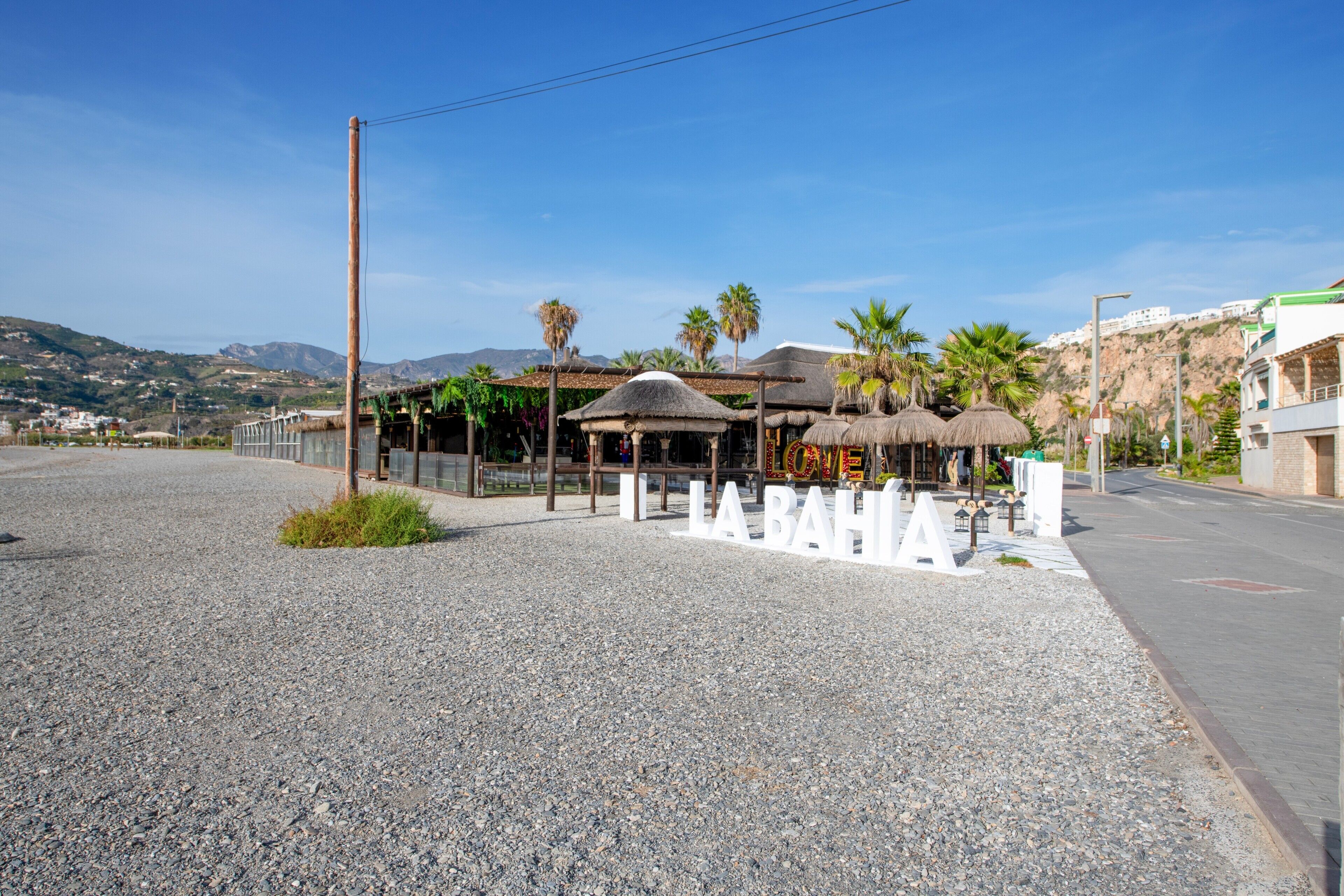 A coastal area with a gravel road, a thatched-roof structure, and a sign reading 'Lone La Bahía.'