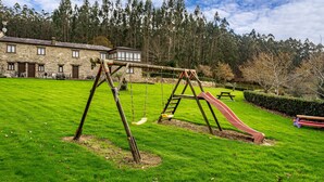 Un conjunto de columpios y un tobogán de madera en un césped, con un edificio de piedra al fondo.
