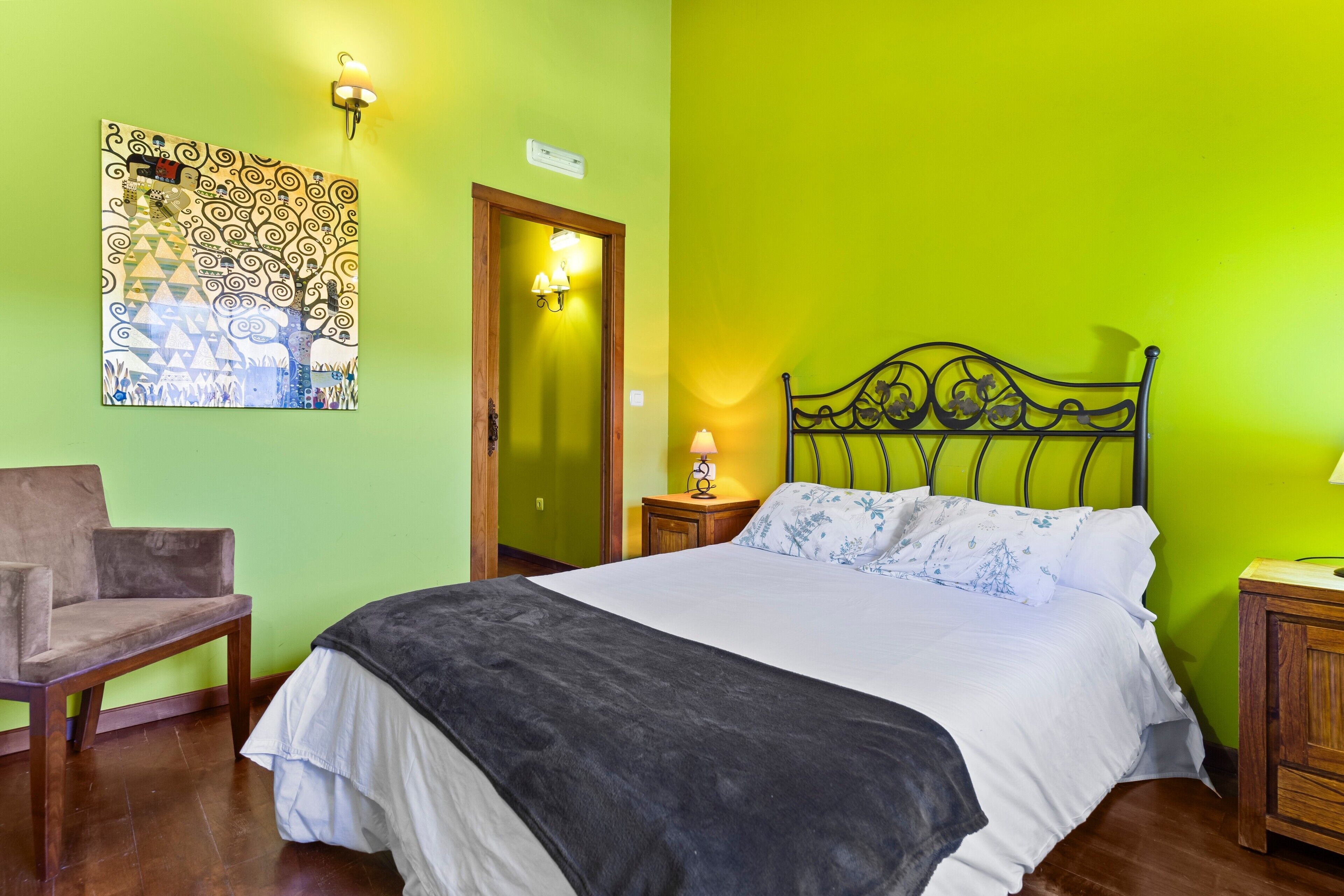 Una habitación con una cama, una silla, un cuadro en la pared, una puerta y una mesita de noche con una lámpara.