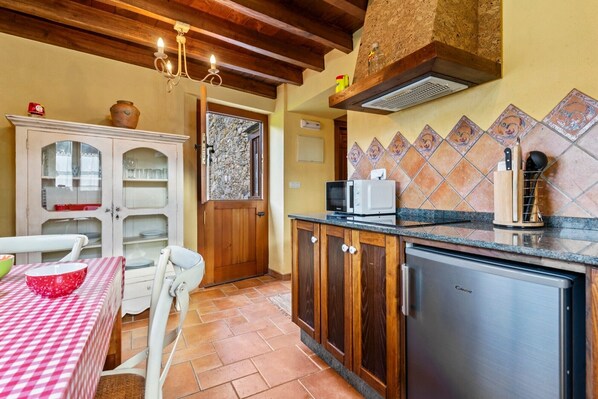A compact kitchen with a microwave, a sink, and a refrigerator. There is a dining table with a checkered tablecloth and a chair. The kitchen has wooden cabinets and a tiled backsplash.