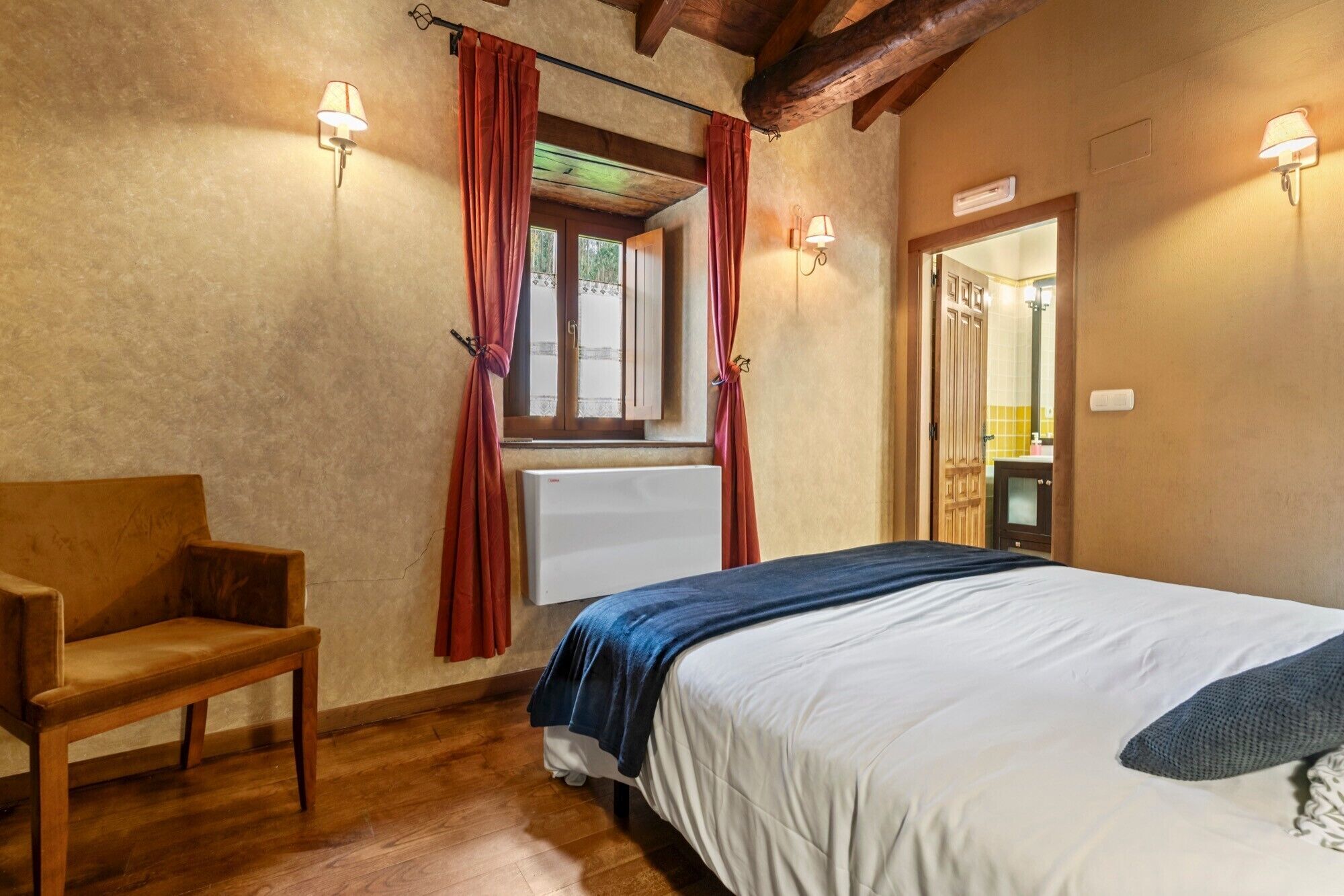 Ein Schlafzimmer mit Bett, Sessel, einem Fenster mit Vorhängen und Wandleuchten.