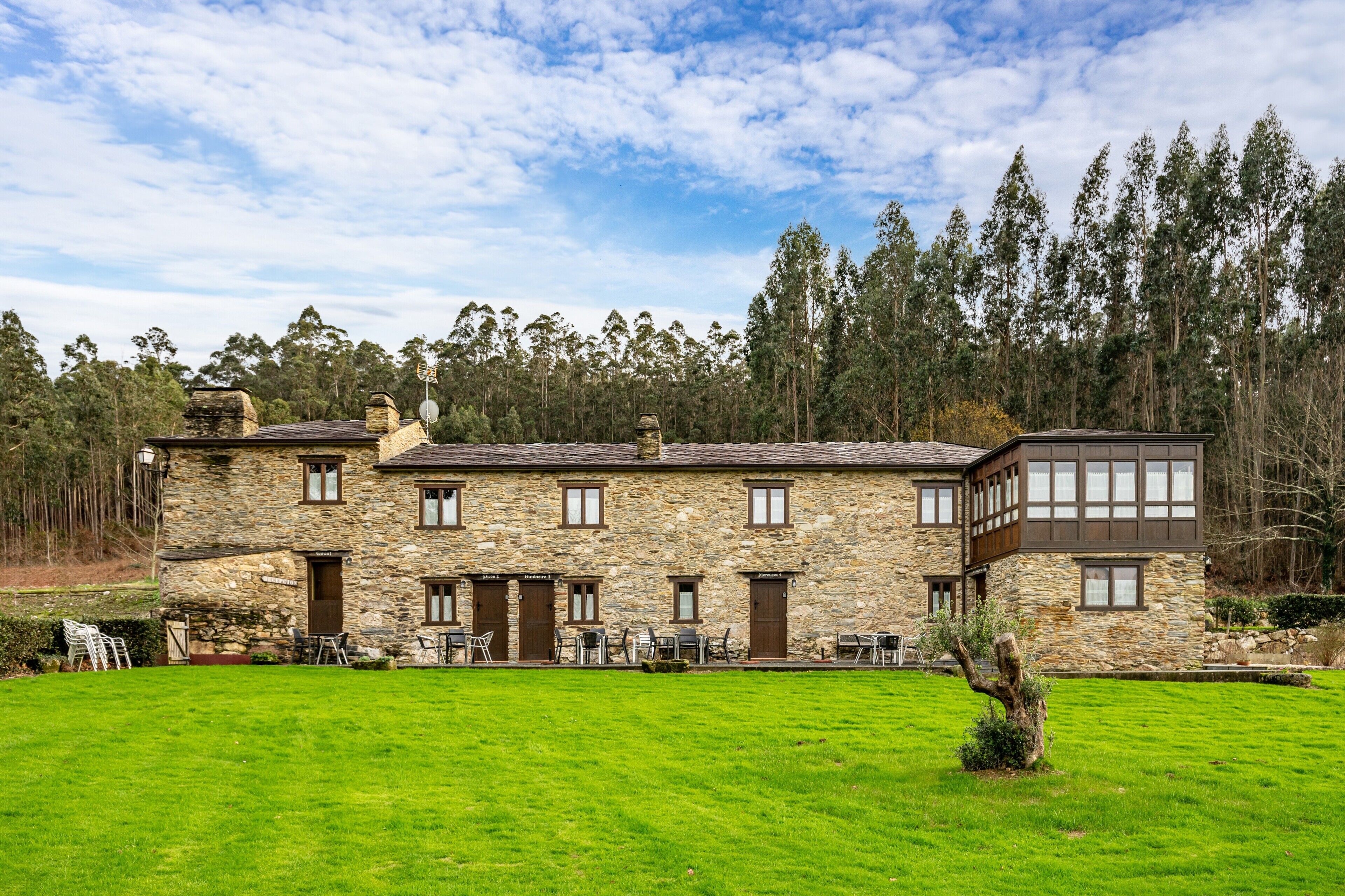 Un edificio de piedra con varias puertas y ventanas, un jardín césped y un bosque al fondo.