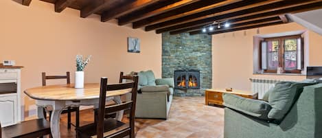 A cozy living room with a stone fireplace, wooden beams, a dining table with chairs, and a green sofa.