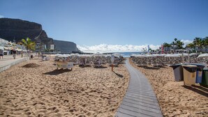 Una playa con un paseo de madera, sillas de playa, sombrillas y un acantilado al fondo.