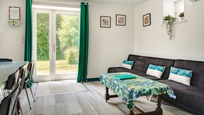 A room with a dining table, chairs, a sofa with cushions, a bed with a floral bedspread, a window with a plant, and a wall-mounted air conditioning unit. - Holiday Home 'Du Parc' with Shared Pool, Lake View & Wi-Fi (Genouillé)