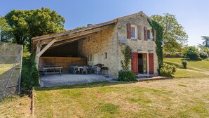 A stone building with a sloped roof, a covered outdoor seating area, and a fenced yard. - Holiday Home 'Du Parc' with Shared Pool, Lake View & Wi-Fi (Genouillé)