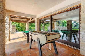 Une table de baby-foot installée dans un espace extérieur abrité, avec des murs en pierre et une vue sur la verdure.