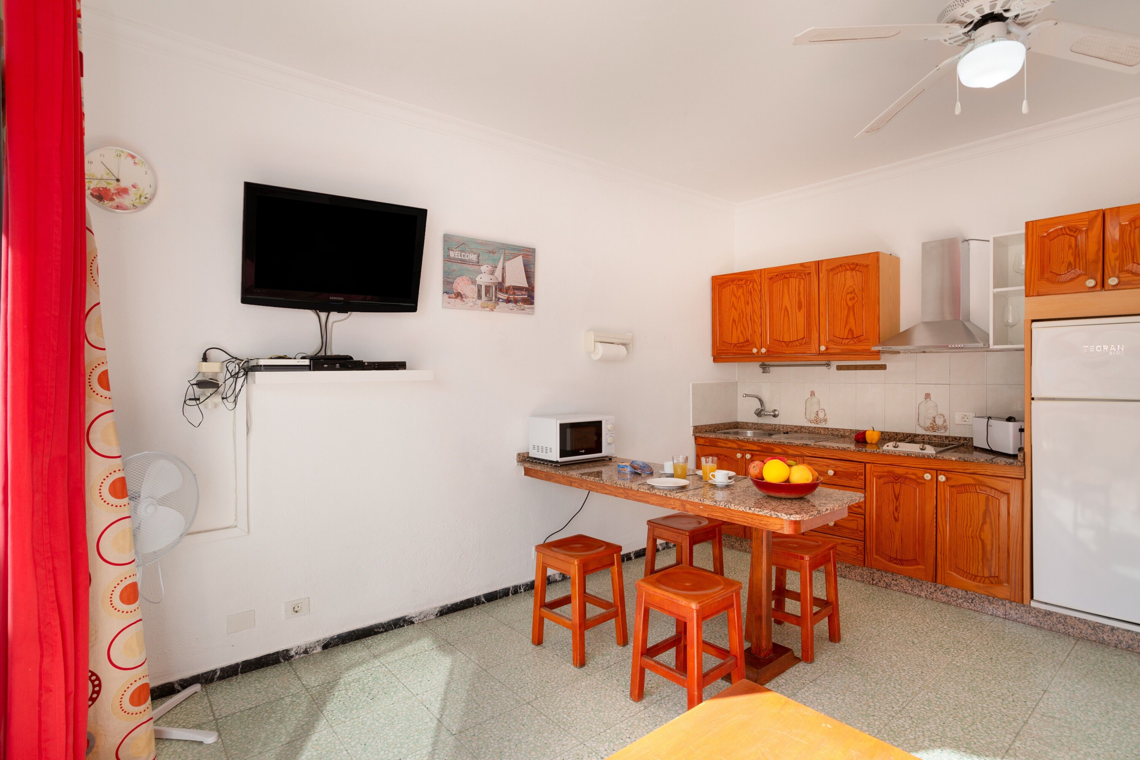 A compact kitchen with a TV, microwave, wooden cabinets, and a dining area with stools.