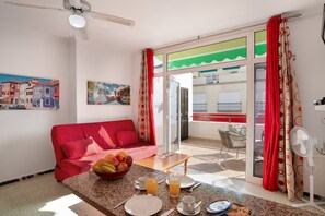 Ein Zimmer mit einem roten Sofa, einem Esstisch mit Frühstücksangebot, einem Ventilator und Blick auf einen Balkon.