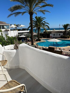 A poolside area with a white wall, a wicker chair, and a palm tree.