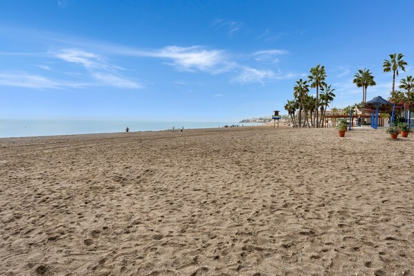 A beach with a clear sky, palm trees, and a structure with a thatched roof.