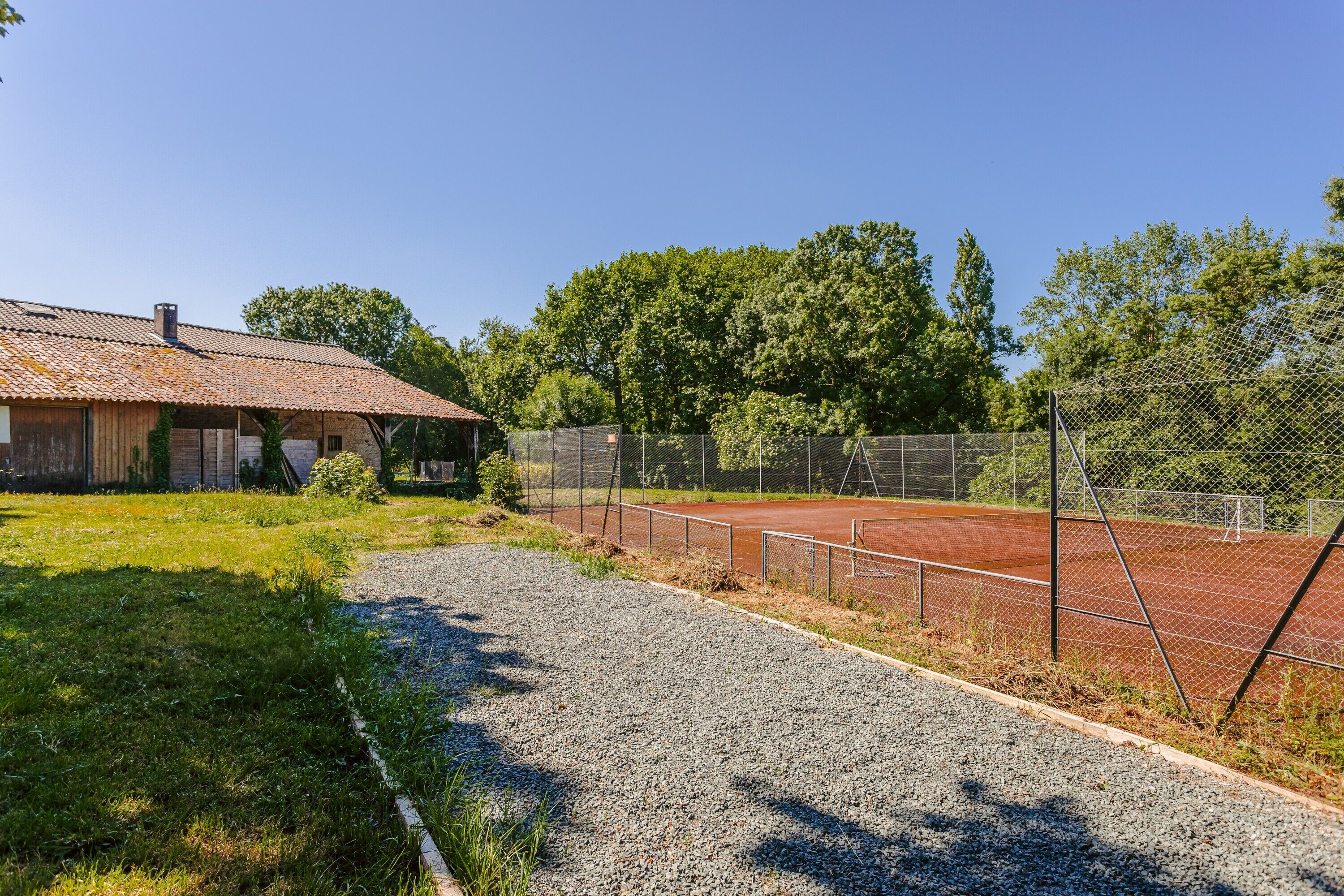 Tenniskenttä, jolta johtaa sorapolkua rakennukseen, jonka katto on päällystetty tiilillä.