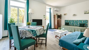 Une pièce comprenant une table à manger, des chaises, un canapé, un téléviseur et une fenêtre avec des rideaux.