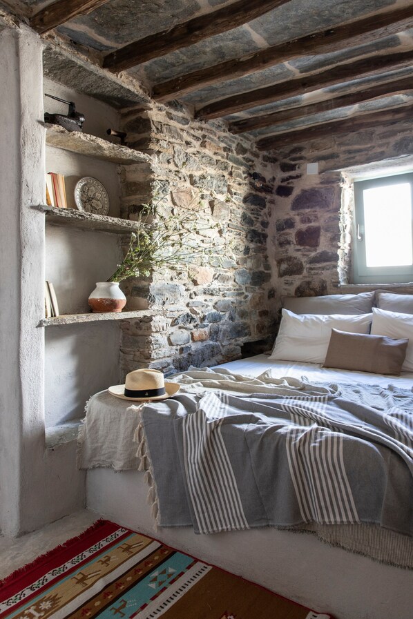 A rustic room with stone walls, a bed with white linens, a wooden beam ceiling, a window, and a shelf with a clock and a vase.
