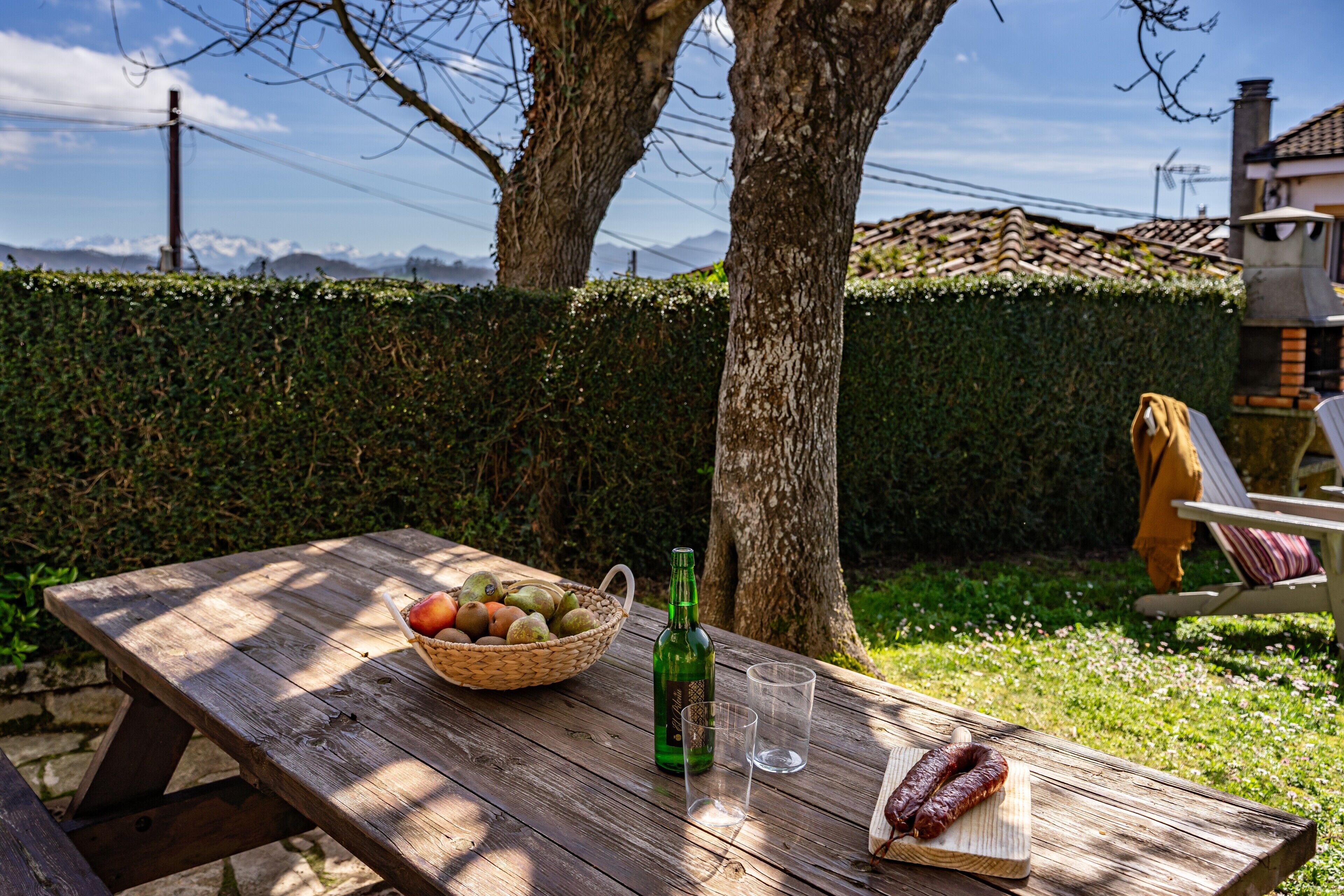 Una mesa de picnic de madera con una cesta de frutas, una botella y salchichas.