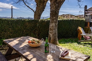 A wooden picnic table with a basket of fruits, a bottle, and sausages.