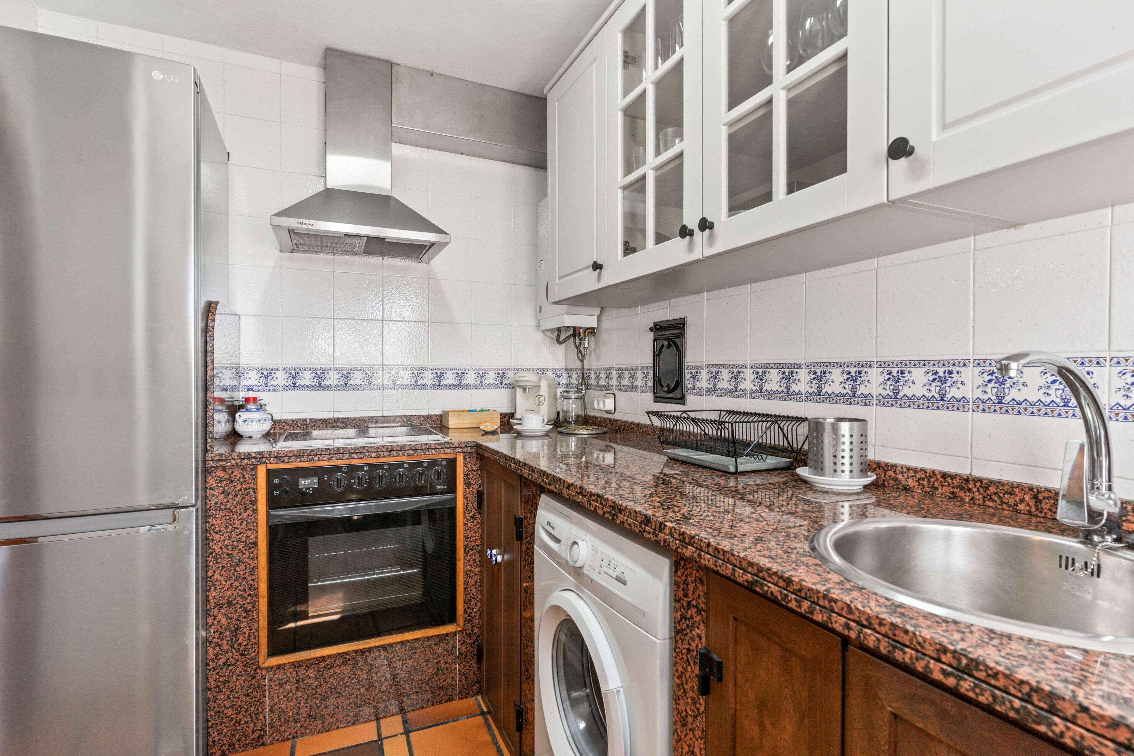 Una cocina con refrigerador de acero inoxidable, horno y lavadora, gabinetes blancos y encimera de granito.