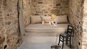 A stone-walled room with a bed, pillows, a hat, a chair, and a pair of sandals. - Apartment 'Onos Eco Living Tinos' with Mountain View, Wi-Fi and Air Conditioning (Falatados)