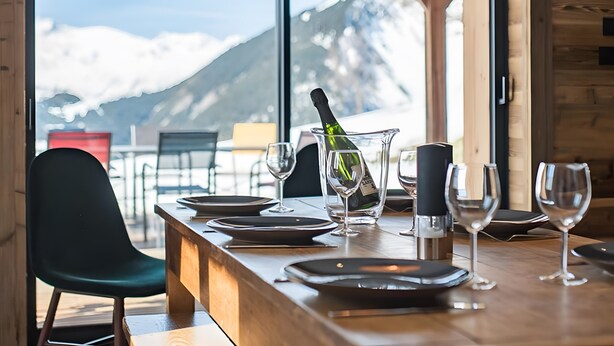 Een gedekte eettafel met borden, glazen en een champagnefles, met uitzicht op een bergpanorama door grote ramen.