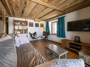 A cozy living room with a sofa, two chairs, a coffee table, a bookshelf, a flat-screen TV, and a window with curtains.