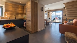 Een moderne keuken met houten kasten, een eethoek met tafel en stoelen, en een woonkamer met een bank en een klein krukje.