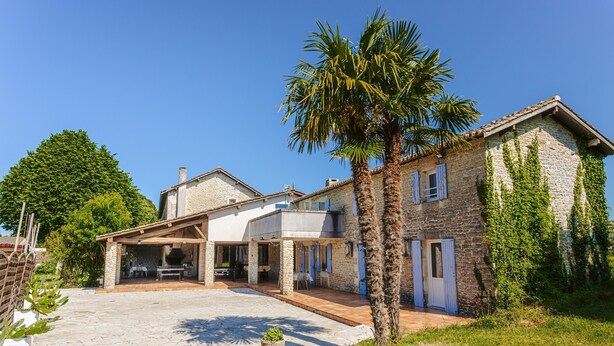 A stone house with a patio, a palm tree, and a large tree in the background.