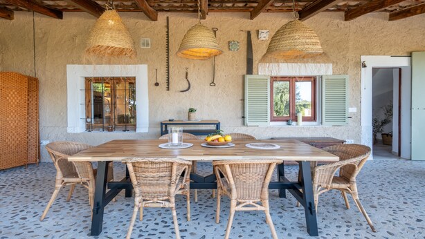A dining area with a wooden table, wicker chairs, hanging lamps, and a rustic wall with a window and shutter. - Finca 'Cas Forner' with Mountain Views, Private Pool and Wi-Fi (Illes Balears)