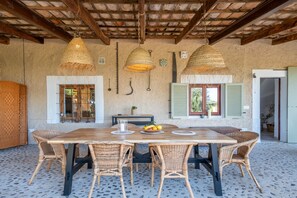 A dining area with a wooden table, wicker chairs, hanging lamps, and a rustic wall with a window and shutter. - Finca 'Cas Forner' with Mountain Views, Private Pool and Wi-Fi (Illes Balears)