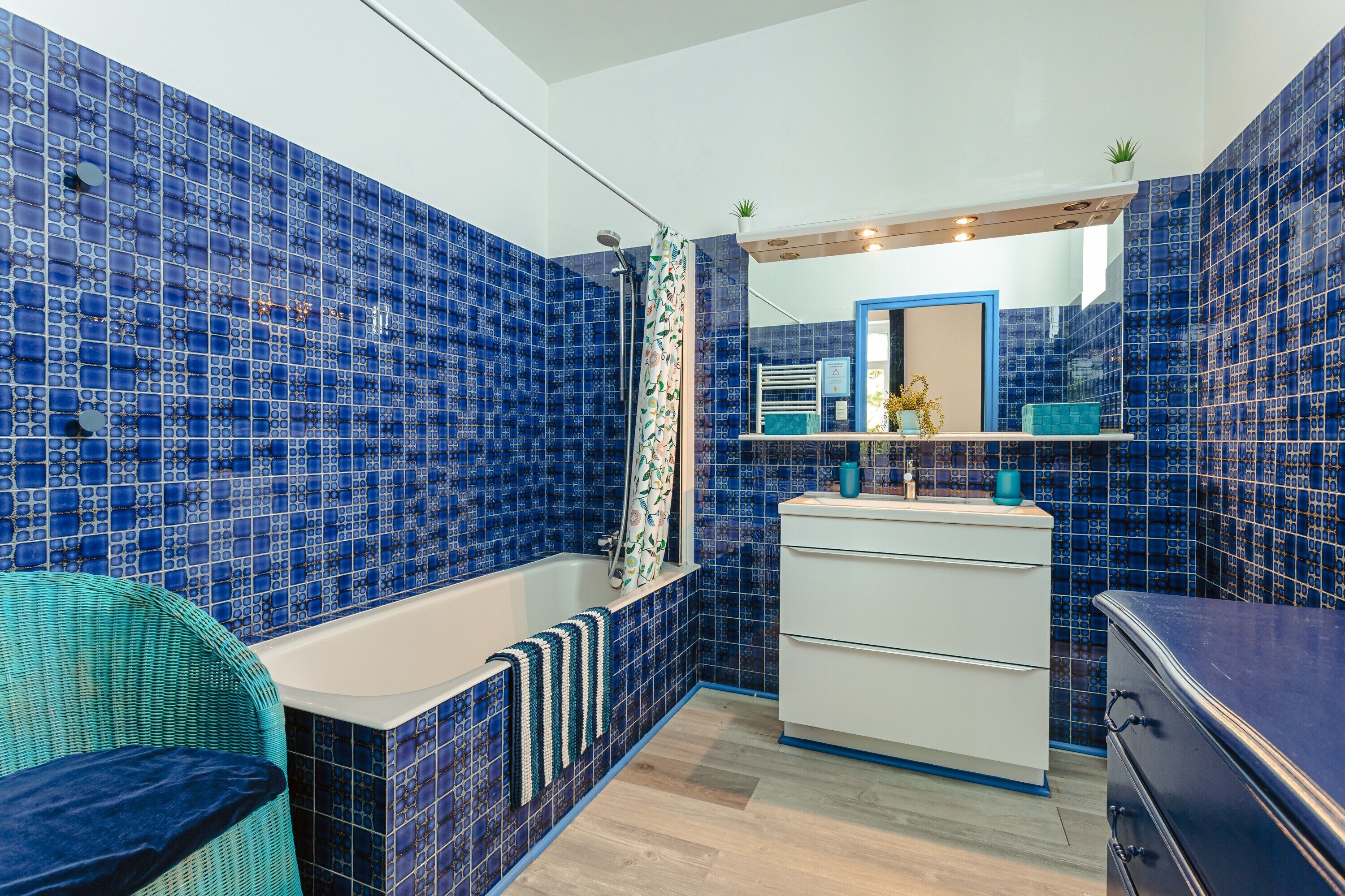 Un baño con mosaicos azules, una bañera blanca, una silla de mimbre, un tocador blanco y un espejo.