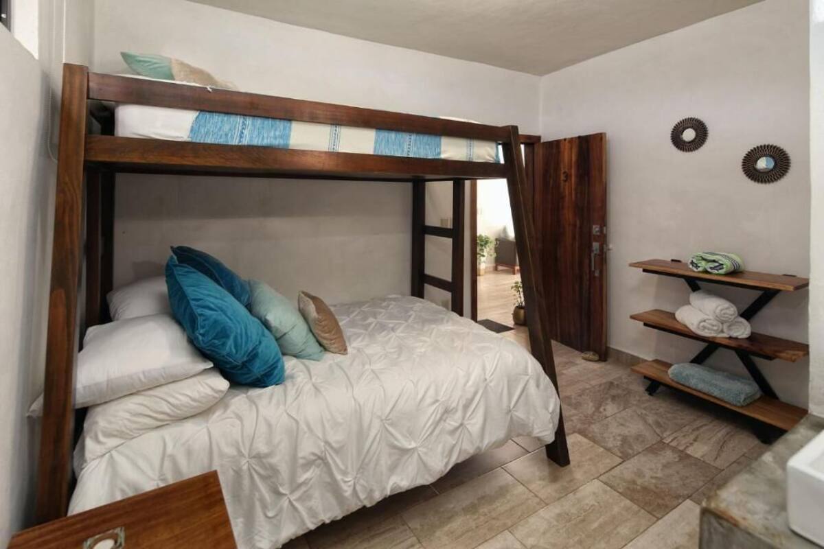 A bunk bed with a wooden frame, white bedding, and blue pillows. There is a shelf with towels and a small plant.