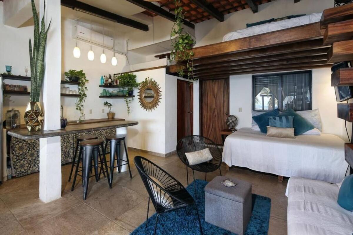 A compact living space with a kitchen area, a dining table with stools, a bed with pillows, and a blue rug.