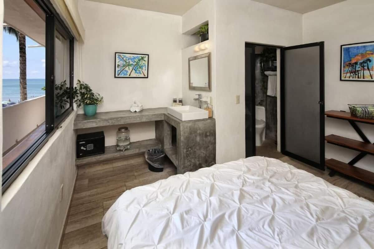 A modern bedroom with a large bed, a built-in vanity with a mirror, a small sink, and a window overlooking the ocean.
